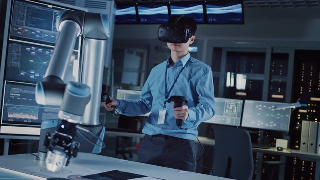 Professional Japanese Development Engineer in Blue Shirt is Controlling a Futuristic Robotic Arm with a Virtual Reality Headset and Joysticks in a High Tech Research Laboratory with Modern Equipment.