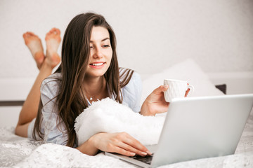 Young smiling woman working on laptop in her bedroom. Happy morning.