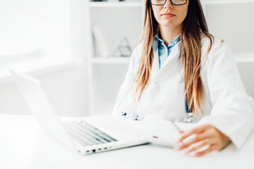 Smiling young female doctor working at the clinic , she is using a computer and writing medical reports.