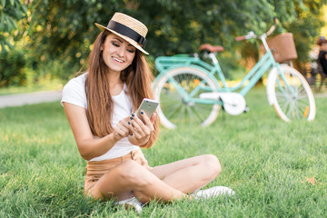  Beautiful young woman sitting on grass and using phone on vintage bicycle background in summer park.