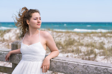 Fototapeta na wymiar Destin, USA Miramar beach city town village day in Florida panhandle gulf of mexico ocean water young woman girl in white dress leaning on wooden fence railing by sand dunes