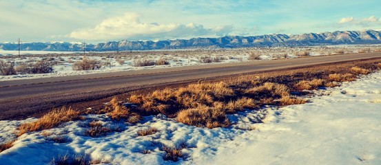 Asphalt Road through snowy field and snow capped mountains in the background.  Taken from rest stop outside Salt Lake City Utah