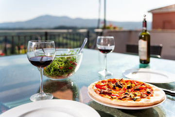 Fresh pizza and salad on glass table terrace outside in Italy with tomato sauce vegetables and two glasses of red wine bottle