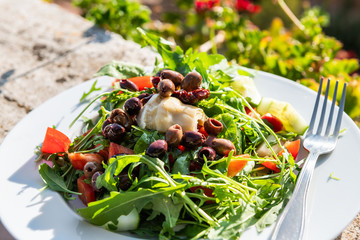 Closeup of arugula salad with olives and artichoke on plate on balcony terrace by red geranium...