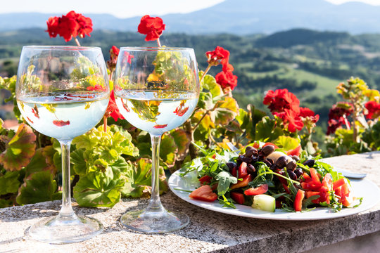 Closeup of arugula salad with olives and white wine two glassses plate on balcony terrace by red geranium flowers in garden outside in Italy in Tuscany