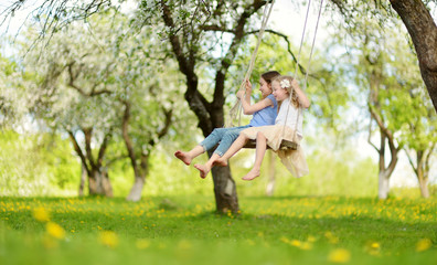 Two cute sisters having fun on a swing in blossoming old apple tree garden outdoors on sunny spring...
