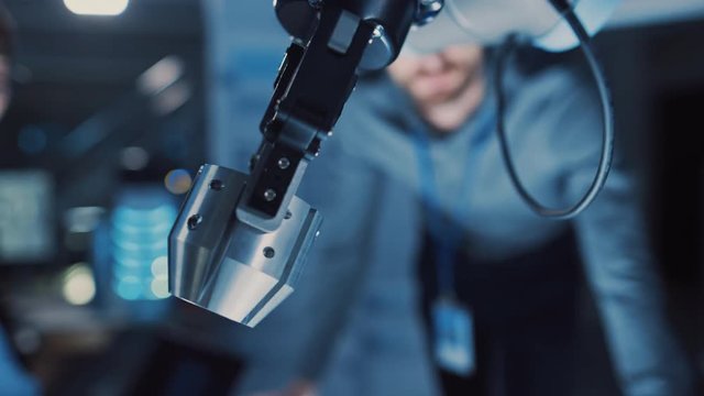 Close Up of a Futuristic Robotic Arm Moving a Metal Object and Placing It. Team of Engineers Observe This Advanced Process. They are in a High Tech Research Laboratory with Modern Equipment.