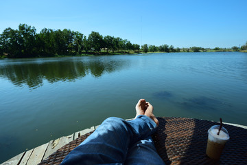 Cropped shot view of man's legs near the lake and a cup of ice coffee, relaxing during summer weekend enjoying good day 