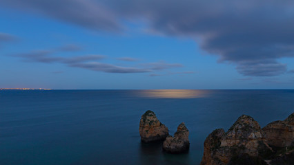 Moonlight shines on a calm surface of Atlantic Ocean. Panorama with distant town at night and horizon over the water, Algarve, Portugal.