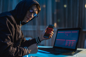 Male hacker in the hood holding the phone in his hands trying to steal access databases. Concept of...
