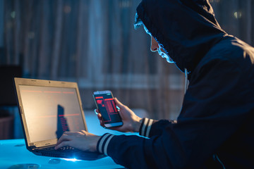 Male hacker in the hood holding the phone in his hands trying to steal access databases with...