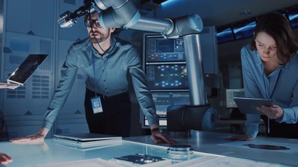 Diverse Team of Engineers with Laptop and a Tablet Analyse and Discuss How a Futuristic Robotic Arm Works and Moves a Metal Object. They are in a High Tech Research Laboratory with Modern Equipment. - Powered by Adobe