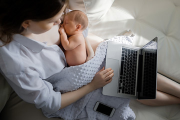 Cheerful young beautiful mother works as a freelancer at home, does remote work, breastfeeds her...