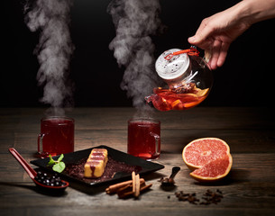 Hand pouring hot tea from glass kettle. Steaming tea in glasses. Sweet rolls in plate, grapefruit, cinnamon sticks, carnation scattered on wooden table, spoon with blueberries on background. Dark view