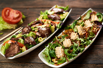 Hot Salads with squid, soybean sprouts, beef, avocado, rucola on recipe of Japanese cuisine in white plate. Chopsticks, tomato, lettuce, basil on wooden table serving in Asian tradition. Top view.