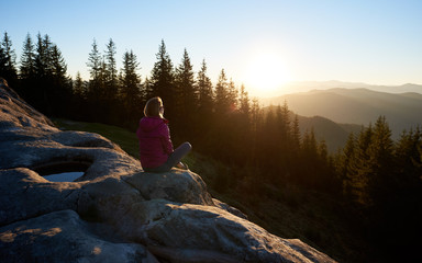 Back view of happy woman hiker sitting on big boulder on the top of mountain in the morning. Female tourist enjoying beautiful sunrise. On background forest, rising sun and blue sky © anatoliy_gleb