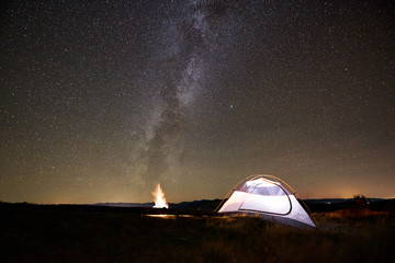 Fototapeta na wymiar Tourist camping at summer night on the top of mountain. Glowing tent beside campfire under amazing night sky full of stars. On background starry sky and mountains. Tourism active lifestyle concept