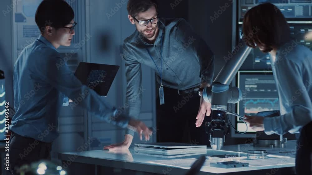 Poster Diverse Team of Engineers with Laptop and a Tablet Analyse and Discuss How a Futuristic Robotic Arm Works and Moves a Metal Object. They are in a High Tech Research Laboratory with Modern Equipment.
