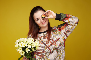 Indoor portrait of  pretty woman with long straight hair, dressed in trendy oversized jumper, closing one eye with cute gentle smile, holding fresh flowers in one hand, colorful Easter egg in another.