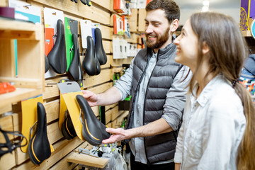 Salesman helping young woman customer to choose saddle standing near the shelves with bicyles parts in the sports shop