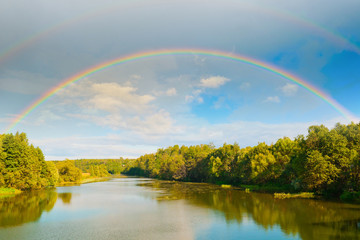 Bright double rainbow in the sky with clouds above the forest and the river