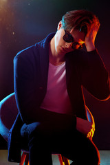 Trendy young man with cool hairstyle wearing black jacket with sunglasses. High Fashion male model in colorful bright neon lights posing on black background. Art design concept