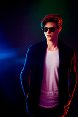 Fototapeta na wymiar Trendy young man with cool hairstyle wearing black jacket with sunglasses. High Fashion male model in colorful bright neon lights posing on black background. Art design concept