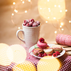 Birthday surprise: french dessert macaroons served on a white plate with fresh raspberries and a mug of fruit smoothie. Background decorated with lights