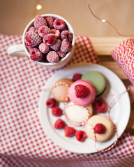 Fresh and sweet dessert: french macaroons with frozen raspberry served on a white plate. Mug of berries, red and white tablecloth. Flat lay