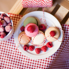 Fresh and sweet dessert: french macaroons with frozen raspberry served on a white plate. Mug of berries, red and white tablecloth. Flat lay