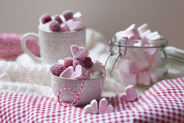 Saint Valentine's day decoration: romantic composition with pink and white marshmallow in the shape of hear, frozen raspberry. Served in ceramic bowl and cup and glass pot. Red tablecloth