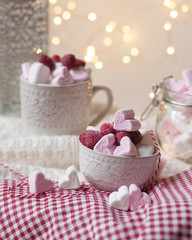 Home saint valentine's day party: heart shaped white and pink marshmallow served with fresh raspberry. Table decorated with red tablecloth and white knitted rud and lights