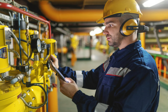 Profile of hardworking worker with helmet, antifones and in protective suit checking air pressure on boilers while standing in heavy industry plant