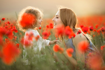 Beautiful smiling child girl with young mother are having fun in field of poppy flowers over sunset...