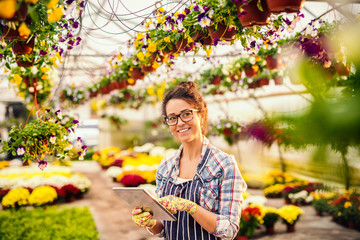 Portrait of beautiful florist with eyeglasses, brown hair and apron while standing in greenhouse. All around flowers.