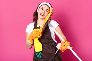 Close up portrait of housewife with good mood, wants to start cleaning her house, has pleasent facial expressions, holds yellow mop and detergent spray oisolated over rose background. Copy space.