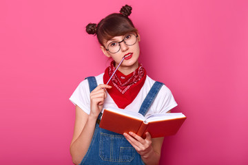 Close up portrait of charming thoughtful student in glasses thinks about something important, holds planner in hands, prepares for exams, keeps pensil at her lip, poses over pink background.