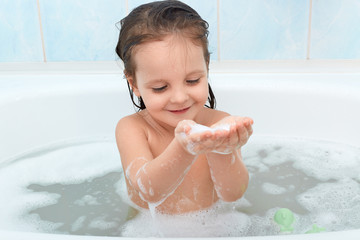 Charming happy baby taking bath, playing with foam bubbles happily. Cute child poses in bathroom on blue background. Infant washing and bathing, relaxes in warm water. Hygiene and health care.