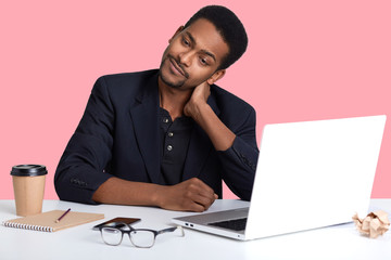 Handsome african american man sitting at desk, wearing classic jacket, looking very tired, putting his hands on his neck, feels pain, looks aside and tries to relax, working online, uses wi fe.