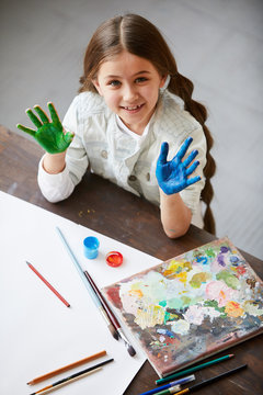High angle portrait of cute little girl showing hands colored with paint and looking at camera, finger painting and creativity concept