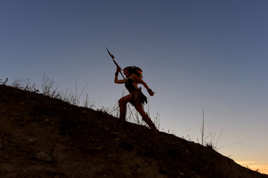 A young girl plays the part of a native American Indian  girl. She poses on top of a hill wit the sun setting behind her.She leaps through the air in an attacking posture.