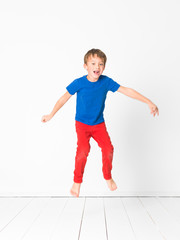 cool, cute boy with blue shirt and red trousers is jumping high in the studio in front of white background and white wooden floor