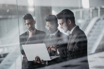 Portrait of three indian co-workers standing with laptop discussing business plan in office