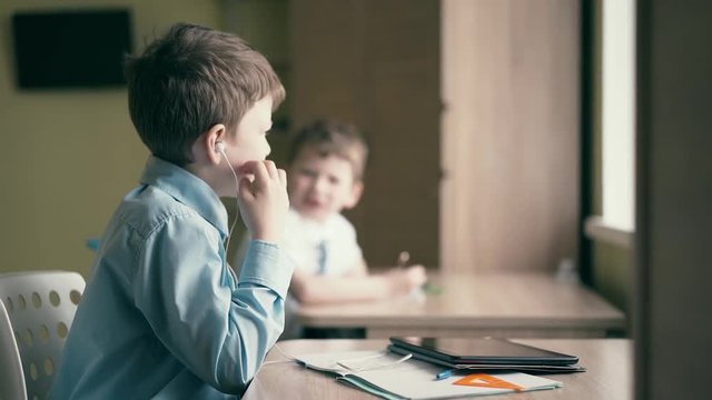 Pupils, little boys, do their homework. One of them makes a drawing in a notebook, the second one sits in headphones and works with a tablet computer.