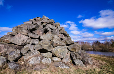 a pyramid made of large, gray boulders; spring in a rural area near a river and a stone heap; stones on a blue sky background