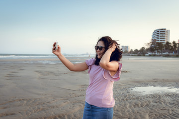 Portrait of Woman is Using Smartphone at The Beach During Sunset, Asian Tourist is Relaxing With Her Cell Phone on The Beach in Holiday Time. Technology Communication Concept.