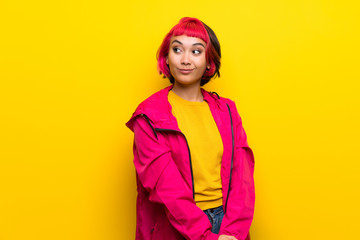 Young woman with pink hair over yellow wall making doubts gesture looking side