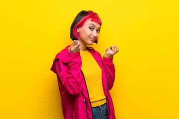 Young woman with pink hair over yellow wall making money gesture