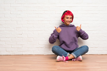 Young woman with pink hair sitting on the floor with thumbs up gesture and smiling