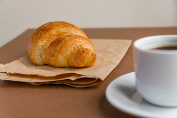 Tasty breackfast. French croissant served on craft paper and cup of black coffee or espresso on brown background. Copy space.